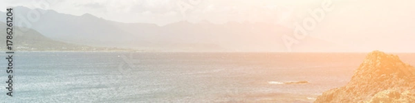 Fototapeta Peaceful coastal landscape with rocky shoreline, calm ocean waves, distant mountain range, and warm sunlight creating a serene natural outdoor atmosphere for travel projects.