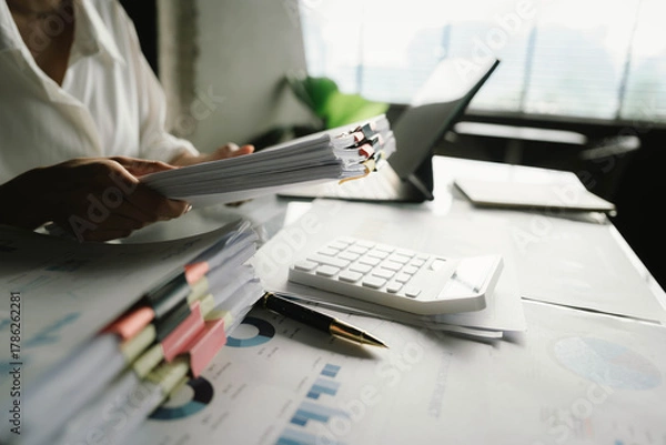 Fototapeta A person working with a calculator and financial documents on a desk, analyzing data from charts displayed on a laptop in a bright office environment.