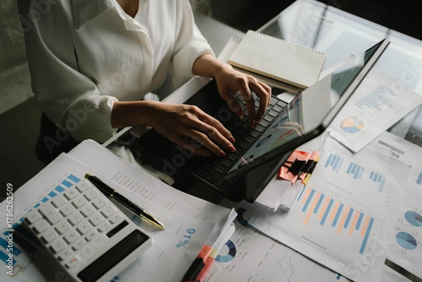 Fototapeta A person working with a calculator and financial documents on a desk, analyzing data from charts displayed on a laptop in a bright office environment.