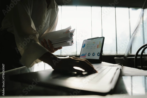 Fototapeta Close-up of businesswoman working at her desk with laptop and stack of documents, focusing on paperwork, reports and financial analysis in a busy office environment.