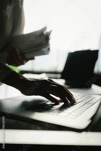 Fototapeta Close-up of businesswoman working at her desk with laptop and stack of documents, focusing on paperwork, reports and financial analysis in a busy office environment.