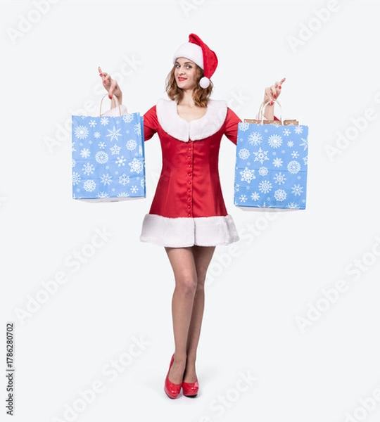 Obraz A cheerful woman dressed in a red Santa outfit and hat holds two blue shopping bags with snowflake patterns, standing on a light background. Festive Christmas shopping and holiday sales concept.