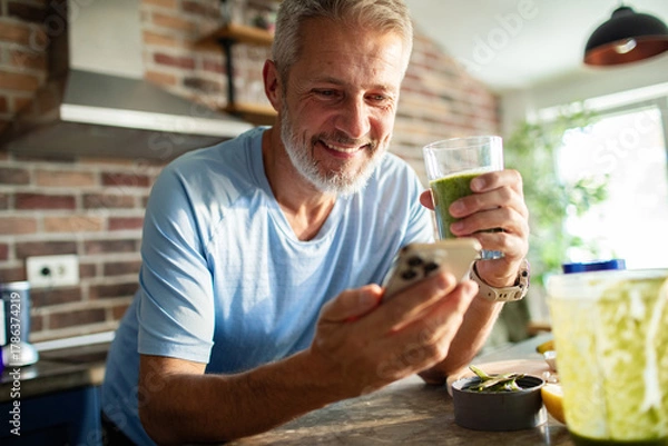 Fototapeta Mature man smiling while drinking smoothie and using smartphone in home kitchen