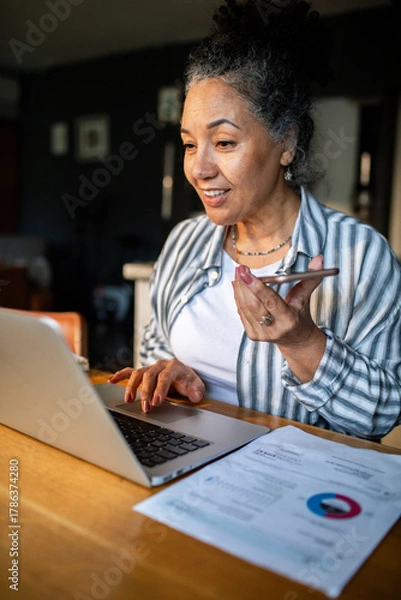 Fototapeta Mature woman smiling, using voice assistant while working at home