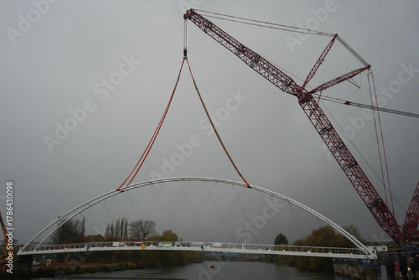 Obraz Massive Red Lattice Crane Carefully Lifts A New White Arched Section Of The Waterside Bridge Over The Trent Basin In Nottingham On A Grey, Overcast Day.