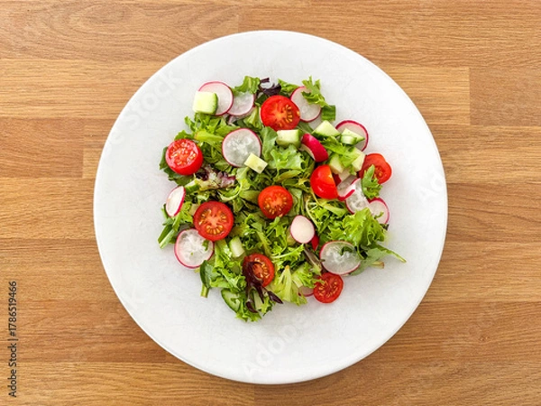 Obraz Simple salad with lettuce, tomatoes, cucumber and radish on a white plate on a wooden table.
