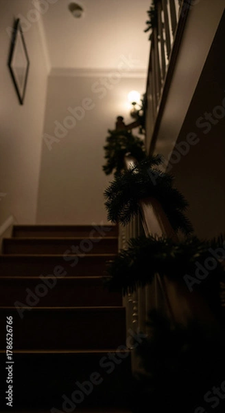 Fototapeta Atmospheric low-angle view of a home staircase decorated with a Christmas garland. A moody, dark scene leading to a warm light, evoking the quiet anticipation and magic of Christmas Eve.