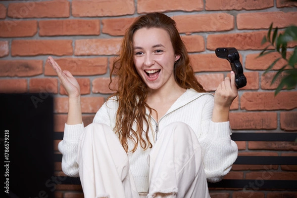 Fototapeta A young beautiful girl sits on the sofa holding a gamepad in her hands and playing a computer game. The player rejoices in victory