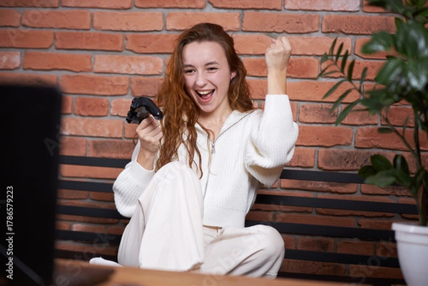 Fototapeta A young beautiful girl sits on the sofa holding a gamepad in her hands and playing a computer game. The player rejoices in victory