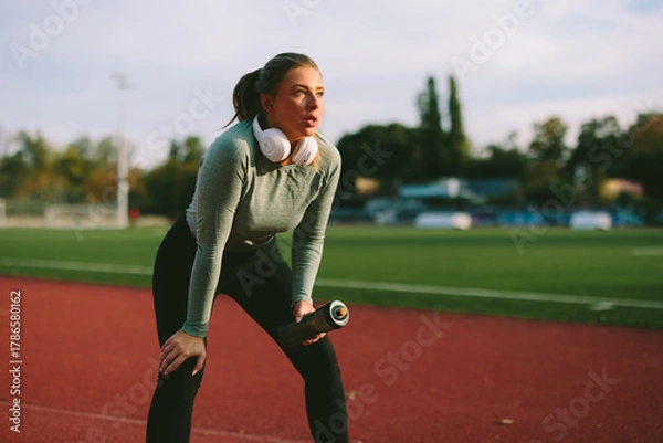 Fototapeta Exhausted but focused Caucasian female athlete leans on her knees to catch her breath on a running track after a strenuous and intense outdoor workout
