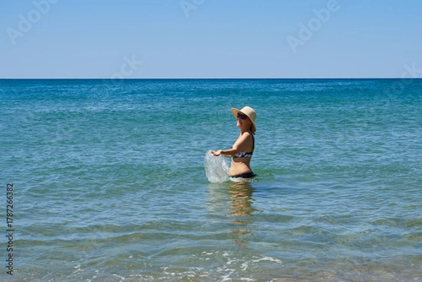 Fototapeta Happy middle-aged woman in a hat splashing water in the sea, enjoying her vacation, the sun
