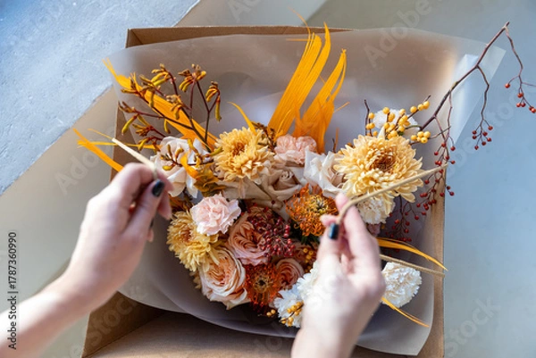 Fototapeta Florist carefully adjusts the elements of a vibrant autumn bouquet, featuring roses and chrysanthemums, inside a craft delivery box