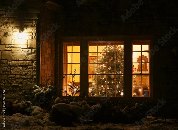 Fototapeta Welcome home Christmas tree.Outdoor close up view of old style brick house window with decorated and glowing christmas tree. Night scene. Christmas and New Year holiday background. 