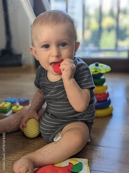Fototapeta A seven-month-old baby sitting on the floor, chewing on a teether toy for gum relief.

