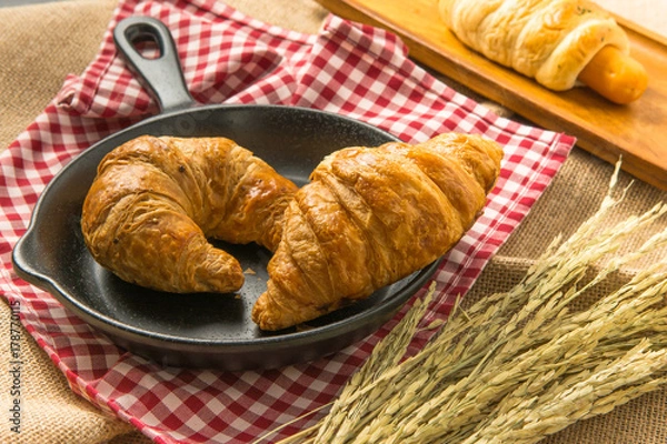 Fototapeta Two pieces of croissant on black ceramic pan that put on the fabric near rice in paddy with sausage filled in bread behind them in yellow sun light for morning meal or breakfast.