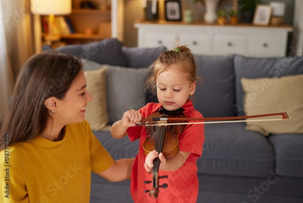 Fototapeta A warm atmosphere fills the living room as a young child experiments with a violin while a parent provides encouragement and guidance. The space is cozy and inviting.