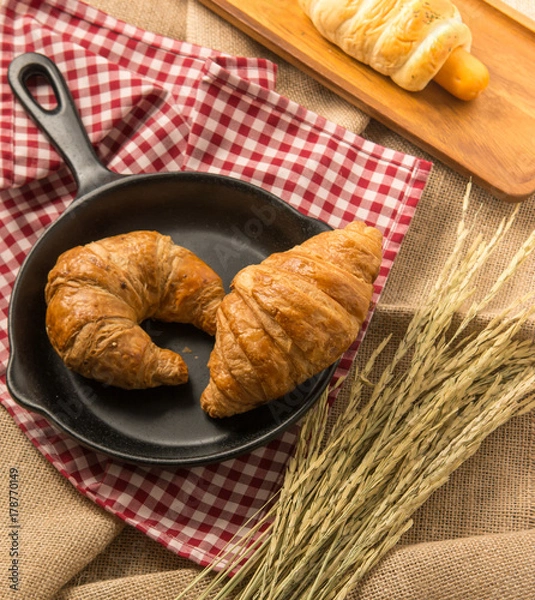 Fototapeta Two pieces of croissant on black ceramic pan that put on the fabric near rice in paddy with sausage filled in bread behind them in yellow sun light for morning meal or breakfast.