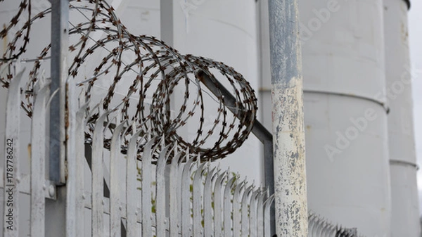 Fototapeta Industrial security scene featuring razor wire along a metal fence and weathered posts near large cylindrical silos, conveying vigilance, containment, and restricted access around an industrial site.