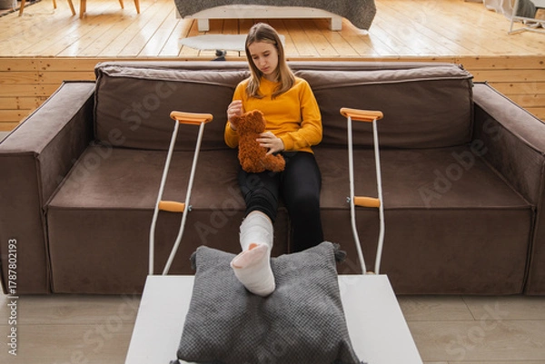 Fototapeta Young girl with broken leg cast sitting on sofa. Sad little kid hugging her teddy bear recovering from broken leg at home. Girl using cast and crutches. Accident injury fracture treatment foot bandage
