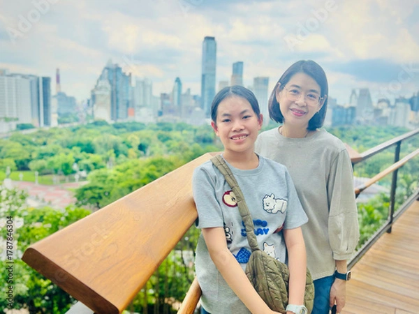 Fototapeta Asian preteen girl and middle-aged mother stand on a rooftop garden in Bangkok, smiling. The city skyline with tall buildings and lush greenery is visible in the background, under a partly cloudy sky