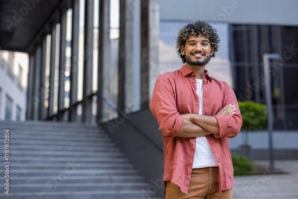 Fototapeta Portrait of a smiling young Indian man in a red shirt standing confidently with his arms crossed on his chest, looking at the camera