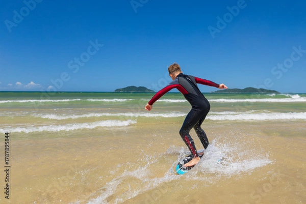 Obraz Young boy skimboarding in shallow water on a sunny beach day. Wearing wetsuit, balancing on board with determination and focus. Perfect image for themes of childhood adventure, active lifestyle, water