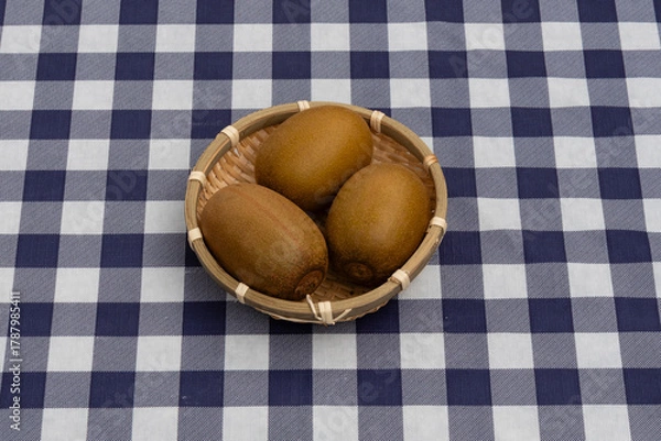 Obraz Top view of three kiwis presented in a small woven bamboo basket on a navy blue and white plaid tablecloth. Ideal for healthy snack and fresh fruit concepts.
