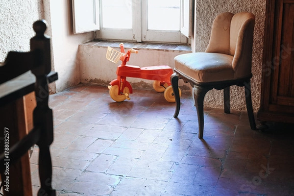 Obraz In a dark room illuminated by soft light from the window stands an old vintage red children’s bicycle. An atmospheric and nostalgic scene filled with warmth and silence.