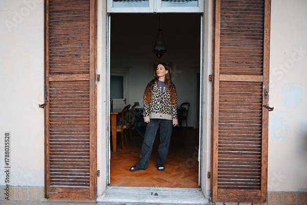 Fototapeta Young woman stands in a doorway, dressed casually in a patterned shirt and jeans, illuminated by soft natural light from the outside.

