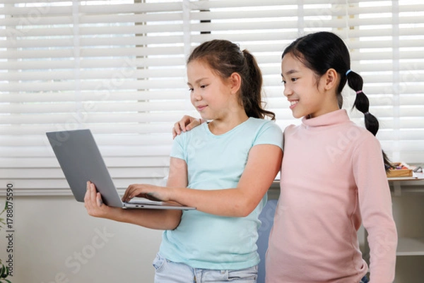 Fototapeta Two girls are standing with laptops in a classroom or study room with a large window. One is typing, while the other smiles and holds her computer,