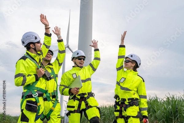 Fototapeta A team of workers in bright yellow safety gear are gathered around a large wind turbine. Group of engineers with PPE, collaborating and walking at wind turbine field, concept of achievement.