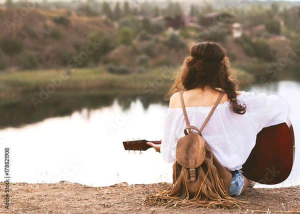 Fototapeta girl in boho style sits on a lake background with a guitar in hand