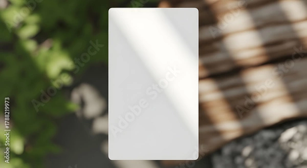 Fototapeta Blank white rectangular box mockup casting a shadow on a textured wooden surface with blurred green foliage in the background