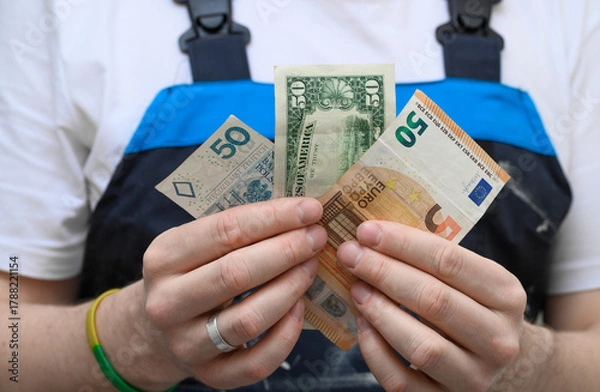 Obraz A handyman holding banknotes in different currencies, euros, dollars and Polish zlotys.