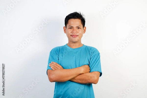 Fototapeta Portrait of a smiling young Asian man in a blue shirt standing confidently with crossed arms. Isolated on white background.