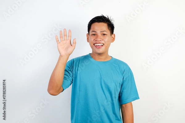 Fototapeta Smiling Young Asian Man Waving Hello in Casual Blue Shirt Studio Portrait for Greeting and Friendly Vibes
