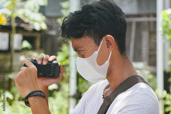 Fototapeta Young Man Filming with Camcorder While Wearing Face Mask Outdoors. Captures modern lifestyle, content creation, and everyday documentation in a natural environment.