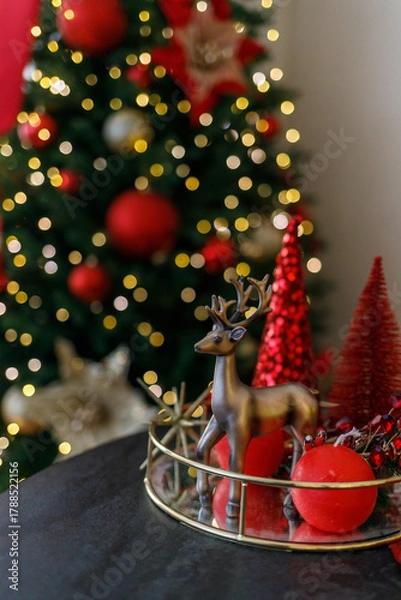 Obraz Beautiful Christmas composition with red decorative trees, a golden reindeer figurine, and shiny gold ornaments on a golden tray. In the background — a glowing Christmas tree with warm fairy lights. 