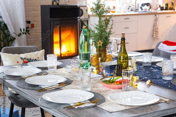 Fototapeta Festive interior of house is decorated for Christmas and New Year in loft style with black stove, fireplace, Christmas tree. Warm studio room with set table, burning wood, cozy and heating of home