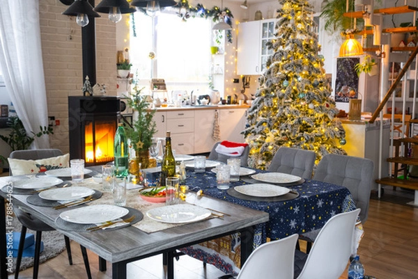 Fototapeta Festive interior of house is decorated for Christmas and New Year in loft style with black stove, fireplace, Christmas tree. Warm studio room with set table, burning wood, cozy and heating of home