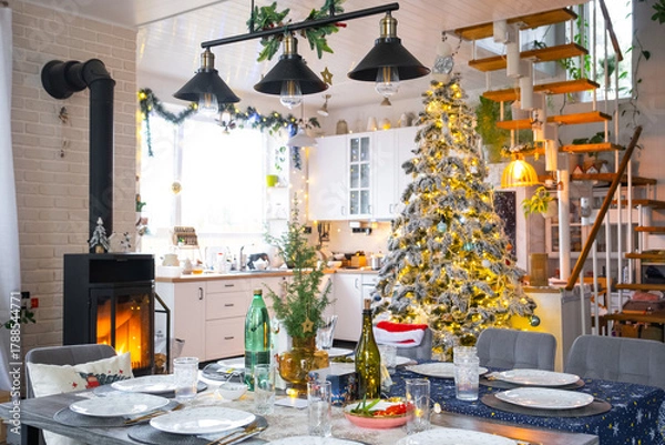 Fototapeta Festive interior of house is decorated for Christmas and New Year in loft style with black stove, fireplace, Christmas tree. Warm studio room with set table, burning wood, cozy and heating of home