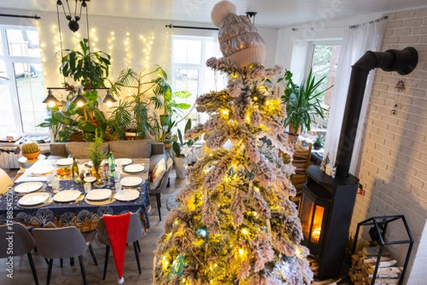 Fototapeta Festive interior of house is decorated for Christmas and New Year in loft style with black stove, fireplace, Christmas tree. Warm studio room with set table, burning wood, cozy and heating of home