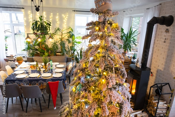 Fototapeta Festive interior of house is decorated for Christmas and New Year in loft style with black stove, fireplace, Christmas tree. Warm studio room with set table, burning wood, cozy and heating of home