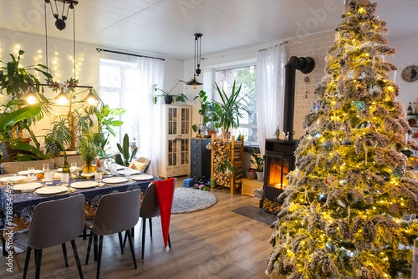 Fototapeta Festive interior of house is decorated for Christmas and New Year in loft style with black stove, fireplace, Christmas tree. Warm studio room with set table, burning wood, cozy and heating of home