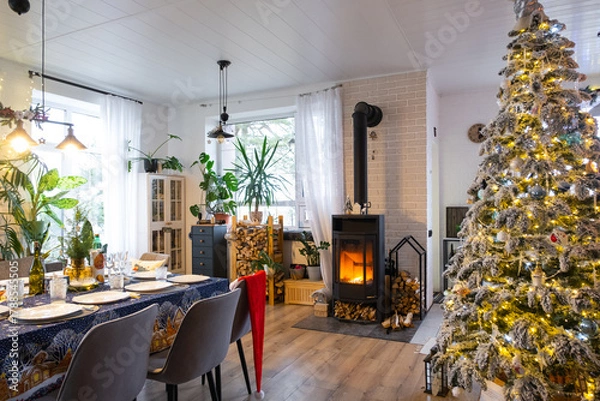 Fototapeta Festive interior of house is decorated for Christmas and New Year in loft style with black stove, fireplace, Christmas tree. Warm studio room with set table, burning wood, cozy and heating of home