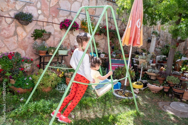 Fototapeta In a colorful backyard garden, two children enjoy swinging on a green swing set surrounded by blooming flowers and plants in terracotta pots. The sunny day brings joy to their playtime.