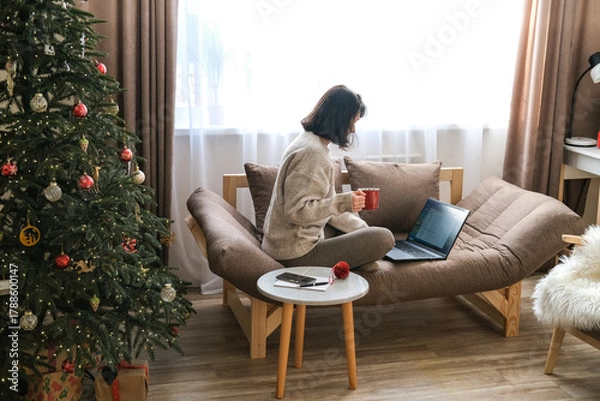 Fototapeta Woman sits on sofa with Christmas tree nearby, working on laptop and holding red mug, creating cozy holiday work-from-home scene. Remote work, online shopping, freelance work during festive period