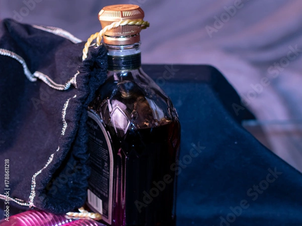 Fototapeta A bottle of brown liquor with a gold top. There is a cloth bag attached to the top. In the lower corner of the photography there are pink small shot glasses. The background is black and grey. 