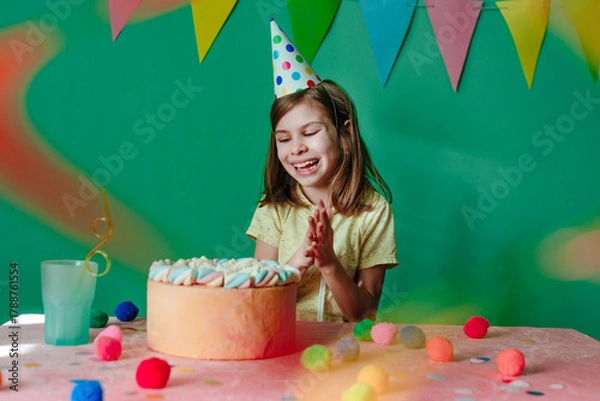 Fototapeta Happy girl wearing party cap sitting at decorated table with cake clapping her hands