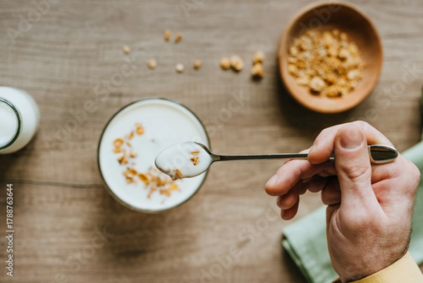 Fototapeta Top view of man adding granola to yogurt bowl on wooden background
